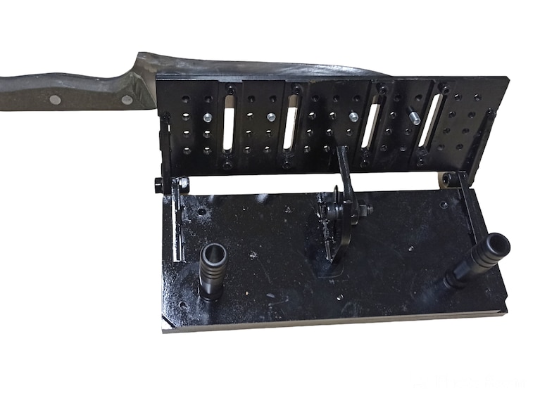 May include: A black metal knife sharpening jig with multiple adjustable slots and a handle. The jig is designed to hold a knife at a consistent angle for sharpening. The handle of the knife is visible on the left side of the image.