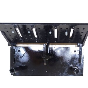 May include: A black metal woodworking jig with a hinged top, featuring multiple holes and slots. The jig has two cylindrical nozzles and is designed for precision work. The overall dimensions are approximately 12 inches long and 8 inches wide.