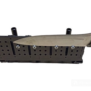 May include: A black knife sharpening system with a stainless steel knife in place. The system has multiple holes and slots, with silver bolts securing the knife. The knife has a green handle and a silver blade.