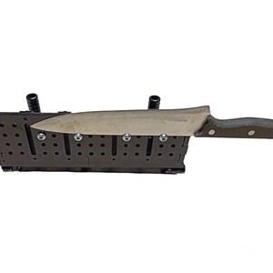May include: A black knife sharpening system with a large chef's knife in place. The system has a series of holes and slots, with the knife blade resting on the system. The knife has a silver blade and a dark green handle.