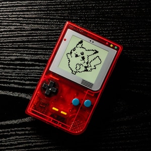 Puede incluir: Una consola de videojuegos portátil roja y gris con una imagen pixelada de Pikachu en la pantalla. La consola tiene un pad direccional, dos botones y una pantalla.