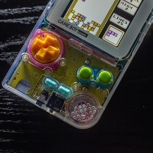 May include: A clear, transparent Game Boy Pocket handheld gaming console with a yellow and green circuit board. The console is displaying a Tetris game on the screen. The screen shows the text "GAME BOY pocket" and "LINES 25".
