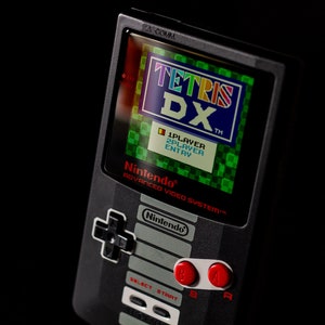 May include: A black and gray handheld video game console with a green screen displaying the text "TETRIS DX" and the words "1PLAYER 2PLAYER ENTRY". The console has a directional pad, four buttons, and two red buttons labeled "L" and "R".