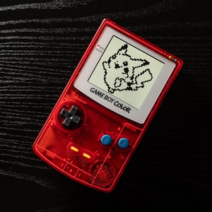 May include: A red Game Boy Color handheld gaming console with a black and white pixelated image of Pikachu on the screen. The console has a blue and white A and B button, a black directional pad, and the text "GAME BOY COLOR" on the front.