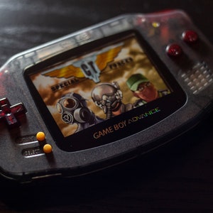 May include: A clear, gray Game Boy Advance handheld gaming console with a red and black design. The screen displays a video game with the text "Special Forces" and "Game Boy Advance".