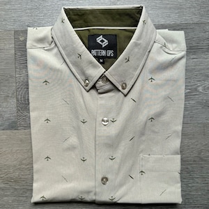May include: A light gray button-down shirt with a green and white airplane pattern. The shirt has a green collar and a label that reads "PATTERN OPS" with the size "M".