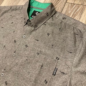 May include: A gray button-down shirt with a green collar. The shirt has a pattern of small footprints, crosses, and airplanes. The shirt has a small label that says "Pattern Legs".
