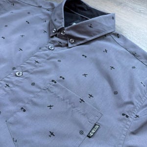 May include: A gray button-down shirt with a small airplane and sunglasses print. The shirt has a pocket on the left side with the word "BITTERFOX" printed on it.