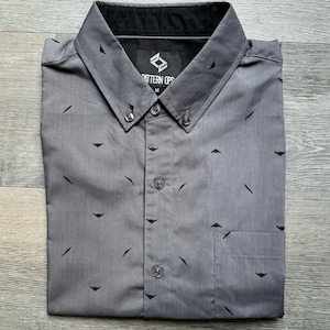 May include: A gray button-down shirt with a black and white pattern of small airplanes. The shirt has a black collar and a pocket on the left side. The shirt has a label that says "Pattern Ops" and "M".