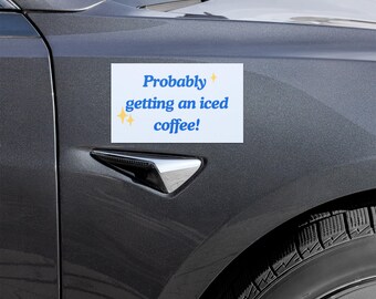 Könnte beinhalten: Ein weißer, rechteckiger Magnet mit dem Text "Probably getting an iced coffee!" in blauer Schrift und gelben Sternchen-Akzenten. Der Magnet ist an der Seite eines dunkelgrauen Autos befestigt.
