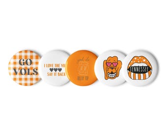 Könnte beinhalten: Fünf runde orange und weiße Knöpfe mit verschiedenen Designs. Der erste Knopf hat ein weiß-orangefarbenes Karomuster mit dem Text "GO VOLS". Der zweite Knopf hat den Text "I LOVE THE VOLS SAY IT BACK" mit drei kleinen Herzen. Der dritte Knopf hat den Text "good ole ROCKY TOP" mit einer Zeichnung eines Bierkruges. Der vierte Knopf hat einen orangefarbenen Cartoon-Hund mit Sonnenbrille. Der fünfte Knopf hat ein rot-weiß gestreiftes Design mit dem Text "TENNESSEE".