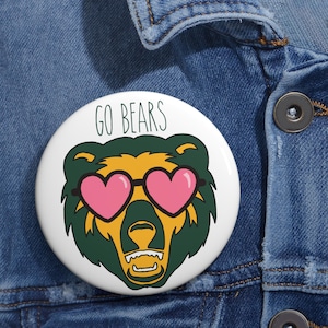 Op de afbeelding: Een witte button met een cartoonbeer met een roze hartvormige zonnebril. De beer is groen en geel omlijnd, met de woorden "GO BEARS" in het groen erboven. De button is vastgemaakt aan een spijkerjasje.