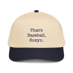Puede incluir: Gorra de béisbol con corona color crema y visera azul marino. El texto "That's Baseball, Suzyn" está impreso en azul marino en la parte delantera de la gorra. La gorra tiene un botón negro en la parte superior.