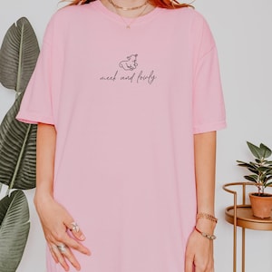 Könnte beinhalten: Ein überdimensioniertes rosa T-Shirt mit einer kleinen schwarzen Illustration eines Lammes und dem Text "meek and lowly" auf der Vorderseite.