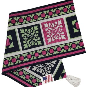 May include: A decorative table runner with a repeating geometric pattern in black, pink, green, and white. The runner features square and diamond shapes with floral accents. A tag with the American flag is attached.