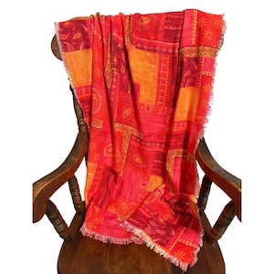 May include: A vibrant, patterned scarf in shades of red and orange, draped over a wooden chair. The scarf features a patchwork design with paisley and floral motifs. The edges are fringed, adding texture to the warm-toned accessory.
