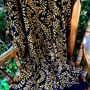 May include: A navy blue fabric with a repeating pattern of gold-colored leaves. The fabric is draped over a wooden rocking chair, with the leaves creating a textured, natural design. The chair's wooden frame is visible.
