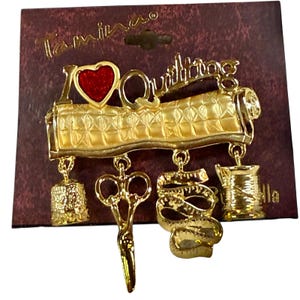 May include: A gold-tone metal brooch featuring the text I Heart Quilting with a red enamel heart. Four charms hang from the base: a thimble, a pair of scissors, a measuring tape, and a spool of thread. The brooch is attached to a dark brown card.