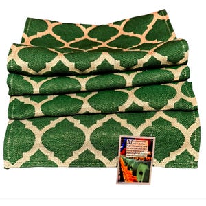 May include: A folded green fabric runner featuring a repeating beige quatrefoil pattern. A small rectangular tag is attached to the bottom right corner, containing text about manufacturing in the Blue Ridge Mountains of North Carolina since 1932.