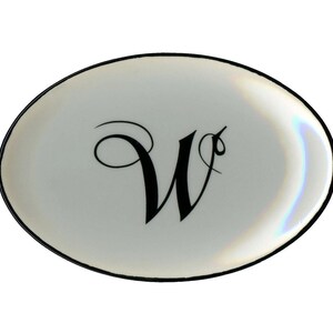 May include: Oval-shaped, white ceramic dish with a black border. The dish features a large, elegant black letter "W" in the center. The surface has a subtle iridescent sheen, adding a touch of elegance.