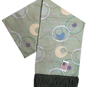 May include: A sage green scarf with a repeating pattern of colorful circles. The scarf has a dark green fringe at the bottom and a tag with the American flag. The circles are outlined in white, pink, and blue.