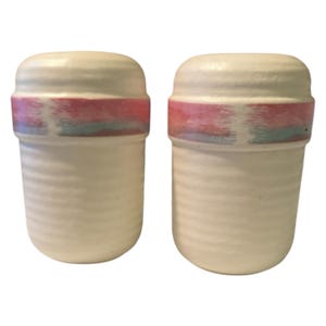 May include: Two off-white ceramic salt and pepper shakers. Each shaker has a cylindrical body with a ridged texture and a rounded top. A band of pink, blue, and purple encircles the middle of each shaker. The shakers are set against a white background.