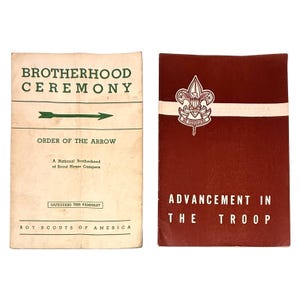 May include: Two vintage Boy Scouts of America pamphlets. The left pamphlet is cream-colored with green text that reads "Brotherhood Ceremony" and "Order of the Arrow." The right pamphlet is maroon with white text that reads "Advancement in the Troop."