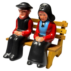 May include: A pair of vintage ceramic salt and pepper shakers depicting a seated couple on a wooden bench. The figures are dressed in traditional Dutch attire, with red jackets, black skirts, and hats. The bench is made of wood.