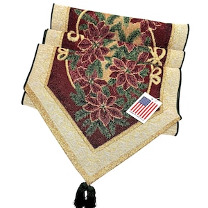 May include: A rectangular table runner featuring a deep red center with a woven poinsettia and green foliage pattern, bordered by a gold textured trim. A black tassel hangs from the pointed end, and a tag displays the American flag and text.