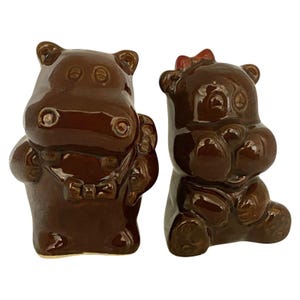 May include: Two brown ceramic hippo figurines. One hippo is standing and wearing a necklace, the other is sitting with a red bow on its head. Both have a glossy finish and are detailed with facial features.
