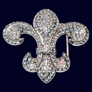 May include: A silver-toned fleur-de-lis brooch, encrusted with numerous clear, sparkling rhinestones. The brooch features a pin closure on the back and is set against a dark blue background. The design is a classic symbol of royalty.