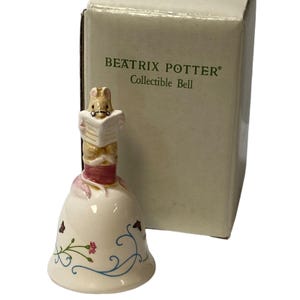 May include: A collectible bell featuring a rabbit reading a book, with a cream-colored bell base adorned with floral and vine designs. The bell is from the Beatrix Potter collection and comes with its original box.