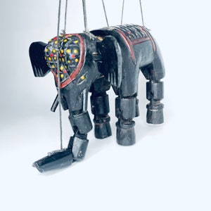 May include: A black wooden elephant puppet with red and yellow painted details. The puppet has jointed legs and a long trunk. It is suspended by strings.