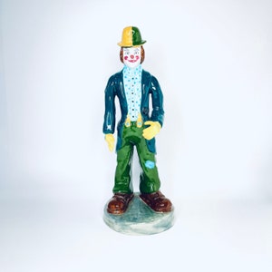 Può includere: Una statuetta di clown in ceramica con un cappello verde e giallo, una camicia a pois blu, pantaloni verdi e scarpe marroni. Il clown sorride e ha un viso dipinto di bianco con labbra rosse e un naso rosso.