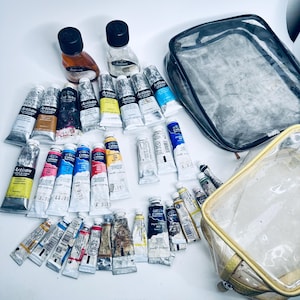 Puede incluir: Una colección de tubos de pintura de artista en varios colores, incluyendo amarillo, azul, rojo y marrón, junto con dos botellas de líquido y dos bolsas de cosméticos transparentes. Los tubos están etiquetados con la marca "Artisan".
