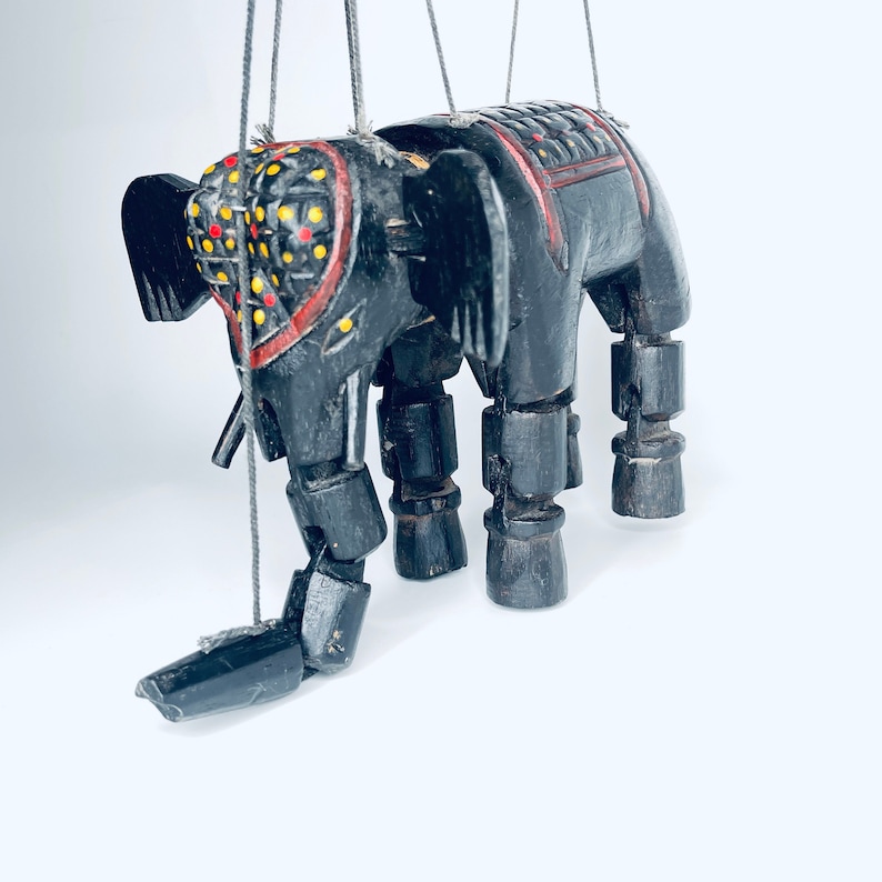 May include: A black wooden elephant puppet with red and yellow painted details. The puppet has jointed legs and a long trunk. It is suspended by strings.