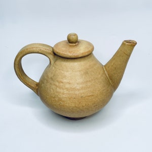 Op de afbeelding: Een lichtbruine theepot van keramiek met een rond lichaam en een deksel. De theepot heeft een handvat en een tuit.