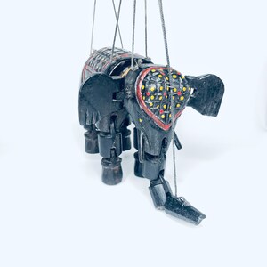 May include: A black wooden elephant puppet with red, yellow, and black painted details. The puppet has strings attached to its limbs and head.