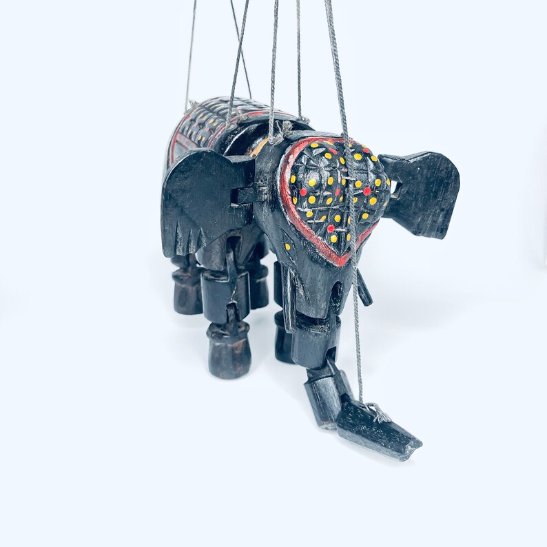 May include: A black wooden elephant puppet with red, yellow, and black painted details. The puppet has strings attached to its limbs and head.