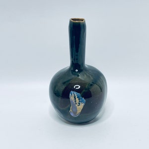 Puede incluir: Un jarrón de cerámica verde oscuro con cuerpo redondo y cuello estrecho. El jarrón tiene un esmalte iridiscente único con toques de morado y dorado.