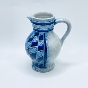 Puede incluir: Jarra de cerámica con asa y boquilla. La jarra presenta un patrón de diamantes azules y blancos en un lado y un diseño azul y blanco liso en el otro. La jarra está sobre un fondo blanco.