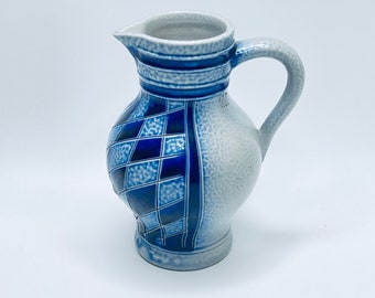 May include: A ceramic pitcher with a handle and spout. The pitcher features a blue and white diamond pattern on one side and a solid blue and white design on the other. The pitcher is on a white background.