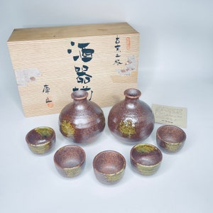 May include: A set of six brown and green ceramic sake bottles and cups in a wooden box. The box has Japanese characters on it. The set is on a white surface.