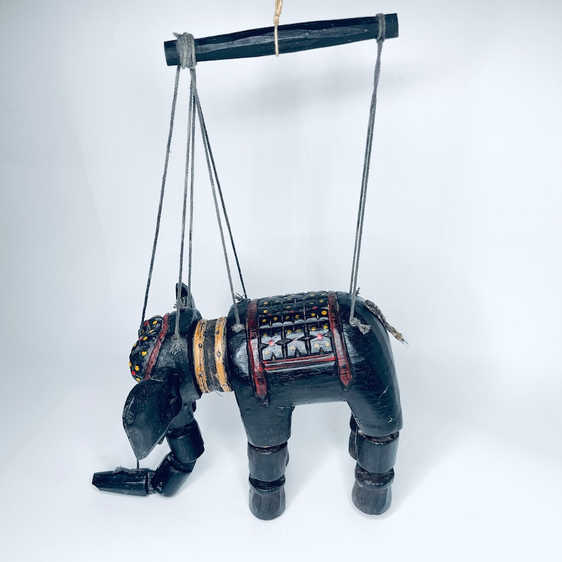 May include: A black wooden elephant puppet with colorful details and a red and black saddle. The puppet is suspended by strings attached to a wooden bar.