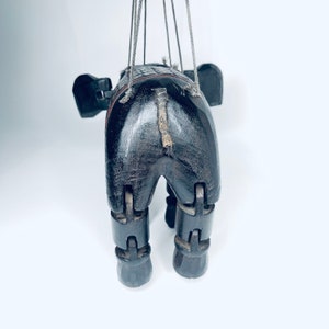 May include: A wooden puppet of an elephant with jointed legs and a tail made of twine. The puppet is painted black and has a red stripe around its middle.