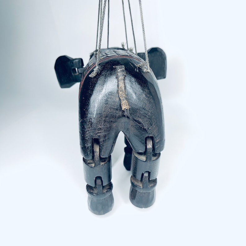 May include: A wooden puppet of an elephant with jointed legs and a tail made of twine. The puppet is painted black and has a red stripe around its middle.