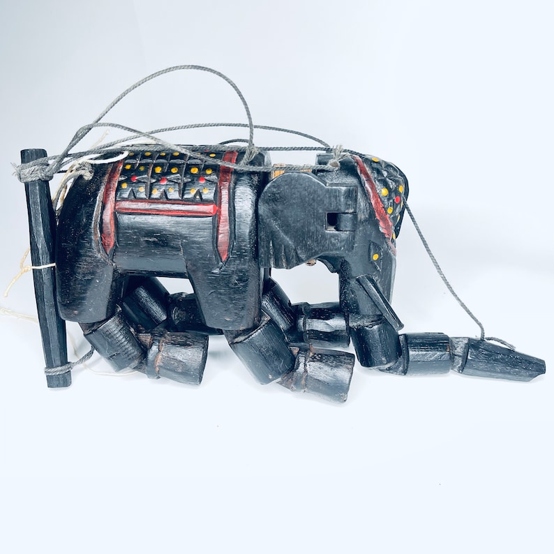 May include: A black wooden puppet of an elephant with red and yellow painted details. The puppet is posed in a kneeling position with its trunk extended. The puppet is attached to strings for manipulation.