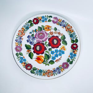 Op de afbeelding: Een wit keramiek bord met een gouden rand heeft een kleurrijk bloemenontwerp. De bloemen zijn rood, roze, oranje, geel, blauw en groen. Het ontwerp doet denken aan traditionele volkskunst.