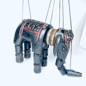 May include: A black wooden puppet of an elephant with red, yellow, and black painted details. The puppet is suspended by strings and has articulated legs and a head.