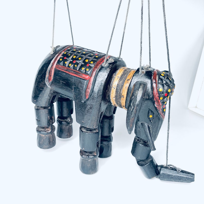 May include: A black wooden puppet of an elephant with red, yellow, and black painted details. The puppet is suspended by strings and has articulated legs and a head.